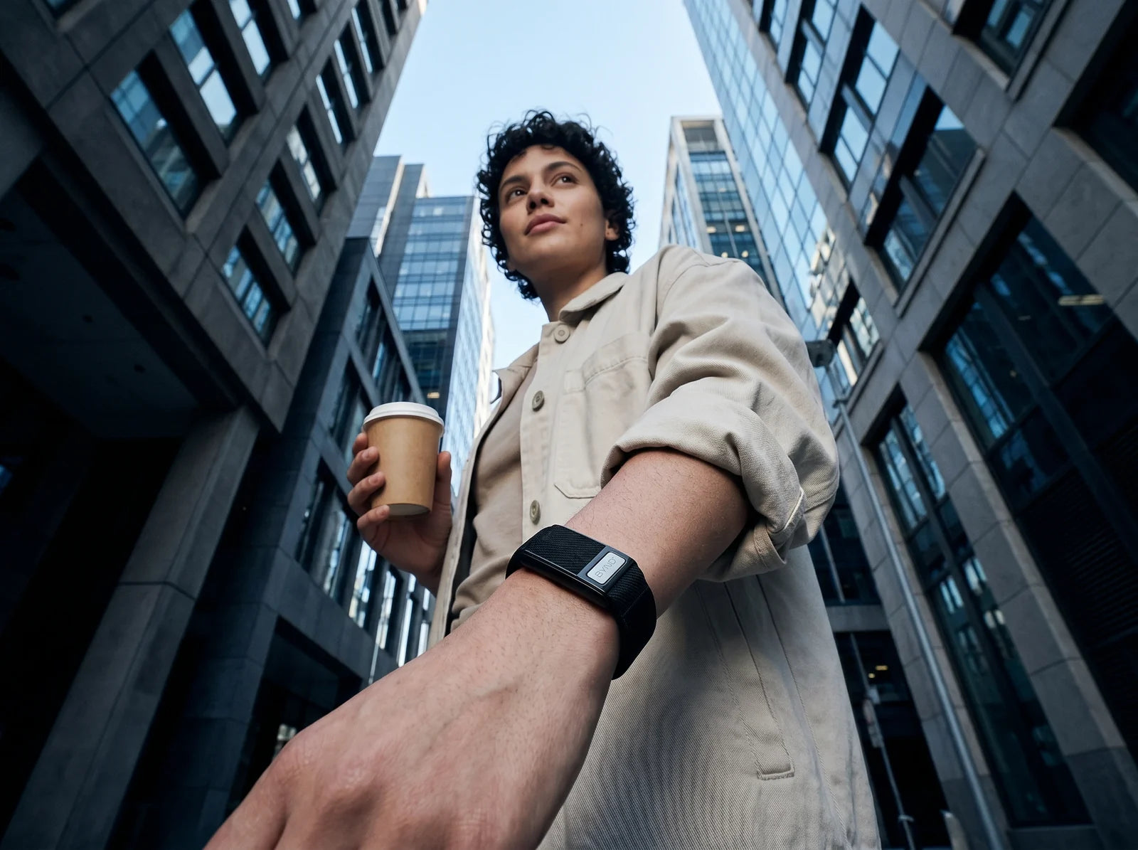 Pessoa com um copo de café entre prédios, vista de baixo, com a BYND no pulso